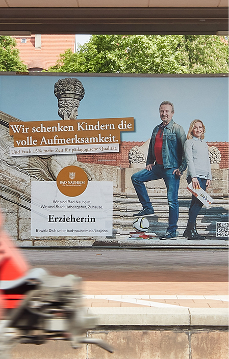 Großflächenplaket der Erzieherkampagne von der Stadt Bad Nauheim hängt am Bahnhof Bad Nauheim, im Vordergrund fährt ein Zug vorbei.
