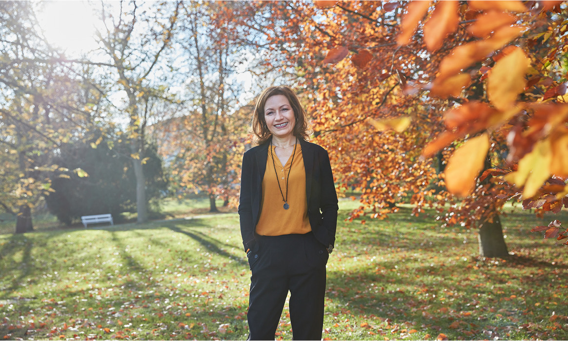 Business-Porträt von Bettina Steeg in einer Parkanlage mit herbstlicher Vegetation und herabfallenden Blättern im Vordergrund.