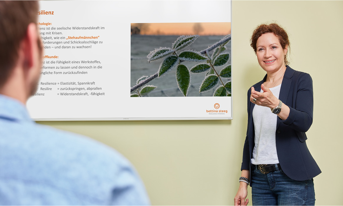 Bettina Steeg während eines Vortrags vor einer Präsentationswand mit einer Person im Vordergrund als Zuhörer.