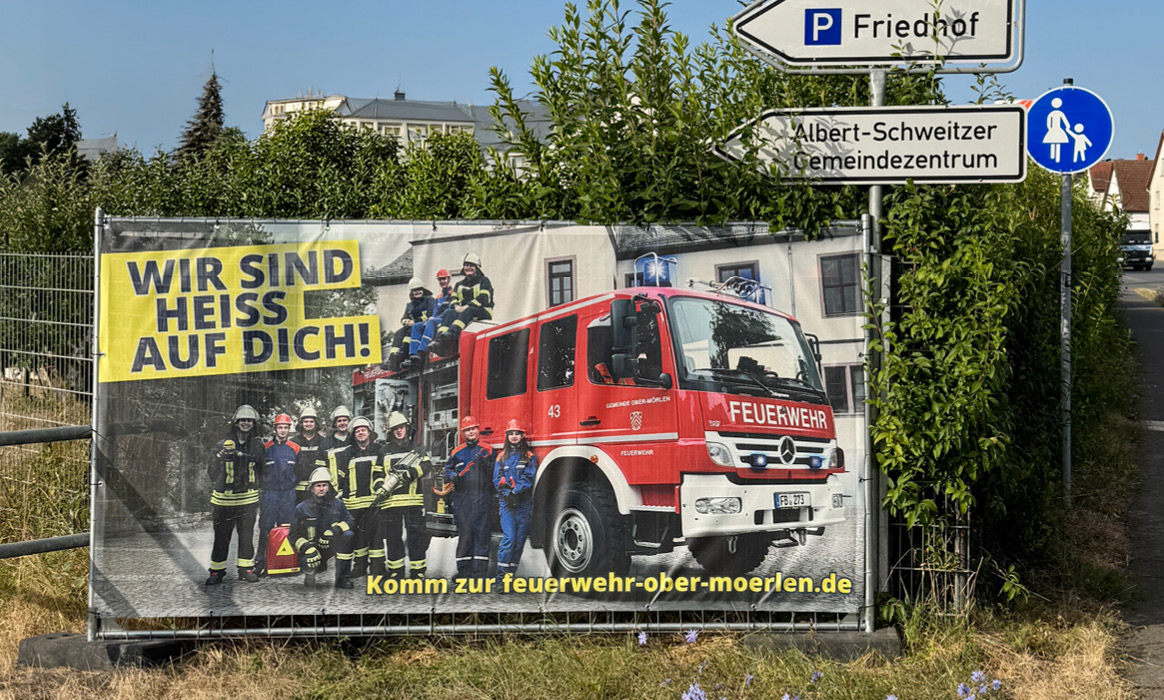 Großflächenbanner der Freiwilligen Feuerwehr Ober-Mörlen mit dem Slogan „WIR SIND HEISS AUF DICH!“, Feuerwehrfahrzeug, Einsatzkräften und Internetadresse, montiert an einem Bauzaun.