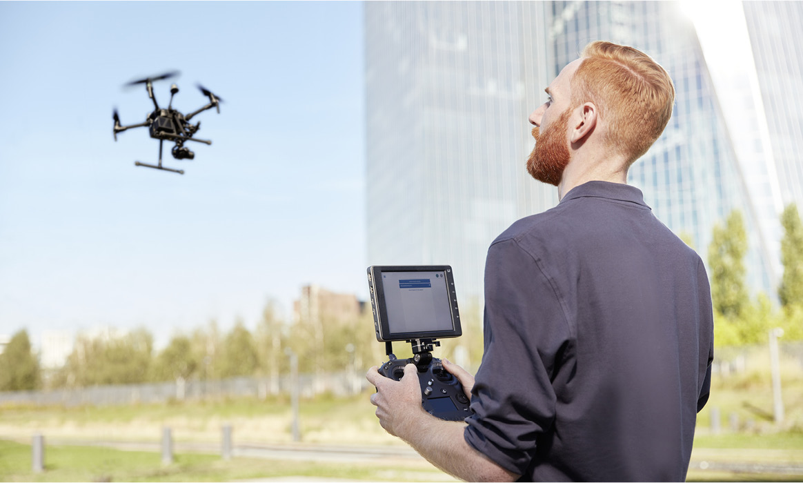 Mitarbeiter von FlyNex steuert eine Drohne mit einer Fernsteuerung vor modernen Bürogebäuden.