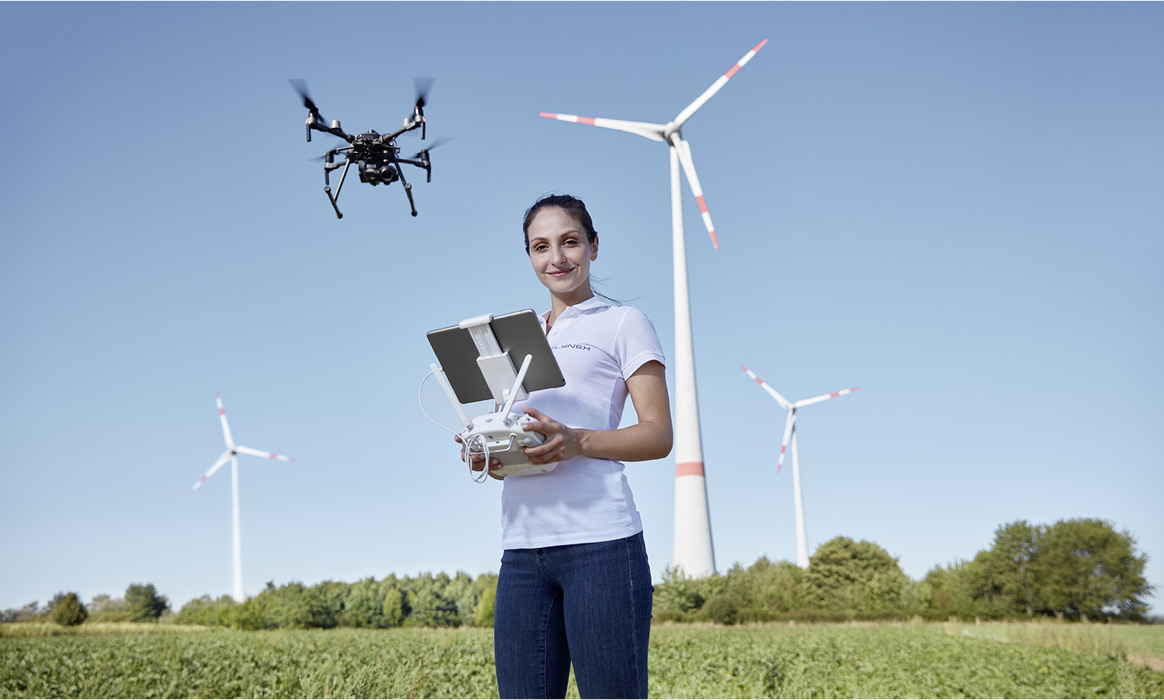 Mitarbeiterin von FlyNex mit Drohnen-Fernsteuerung vor Windkraftanlagen während eines Drohnenflugs.