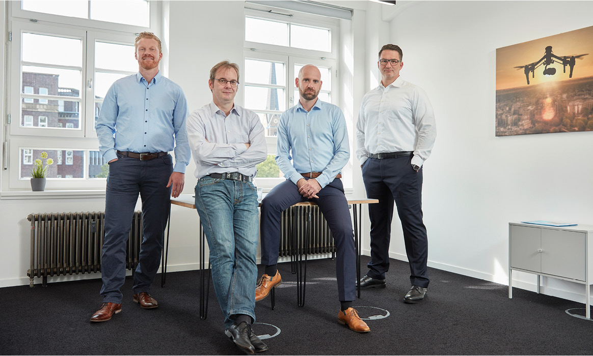 Geschäftsführung von FlyNex als Gruppenporträt in den Büroräumen vor einem Wandmotiv mit einer Drohne.
